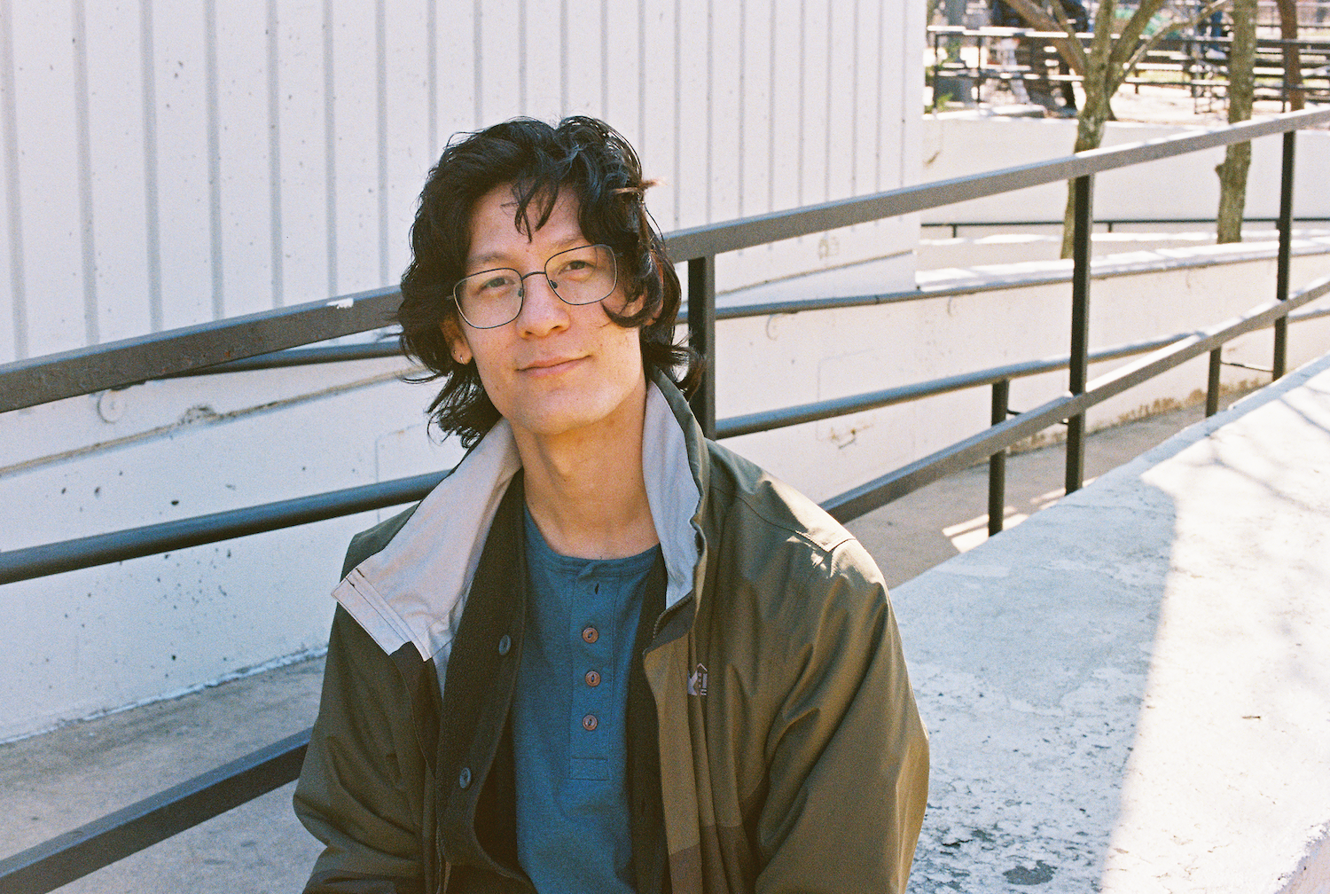 Portrait of Morgan Võ, sitting outside in the sun, wearing long hair and a green rain jacket. They are smiling.