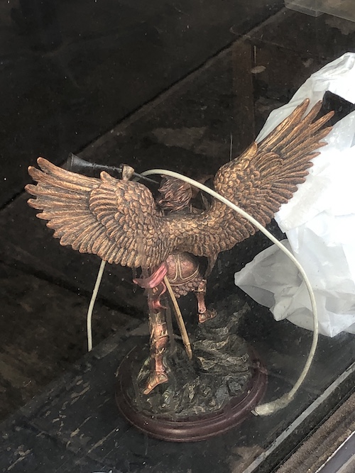 a bronze statuette of an angel with a clarion, around which an iphone charger cable is arcing. photo taken from behind the angel.