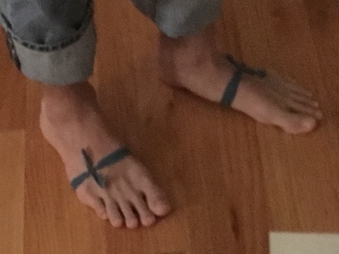 A close cropped digital photograph of Morgan's bare feet on a wooden floor. Each foot is wrapped in a thin strip of forest green cloth, tied with a kind of bow. He also wears jeans cuffed at the ankles.